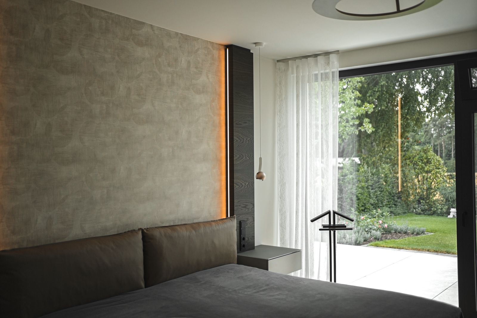 Schlafzimmer mit Polsterbett vor einer strukturierten Wand mit Lichtfuge, daneben ein bodentiefes Fenster zum Garten